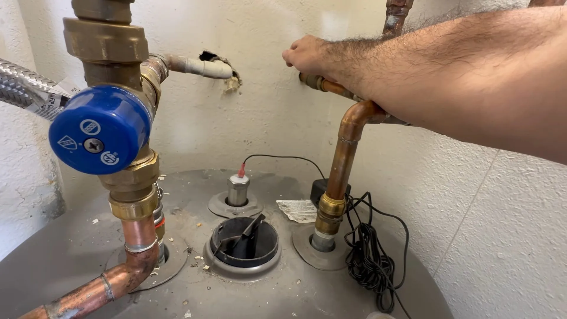 Arm reaching across the top of a gray water heater to a brass fitting beside a blue-handled valve and an anode port