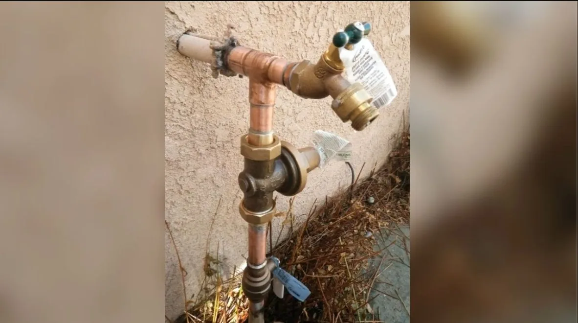 Brass shutoff valve with a paper tag on a copper pipe against a stucco wall, with dry brush at its base