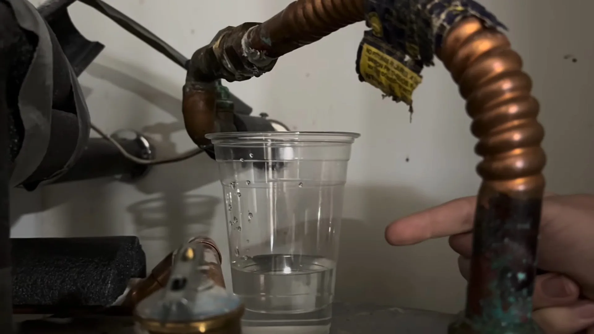 Clear plastic cup held beside a corrugated copper water heater connector with a worn warning tag hanging from the pipe