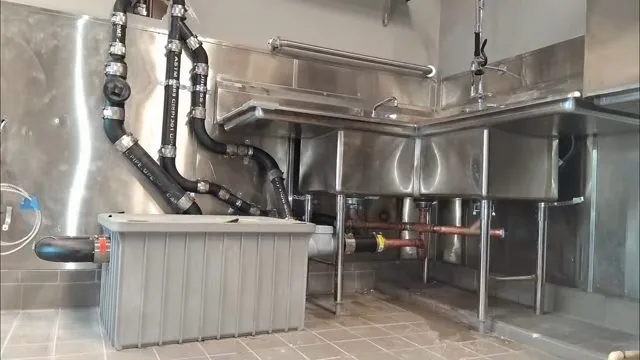 Stainless three-compartment sink in a commercial kitchen with black drain pipes running to a gray grease trap on the floor