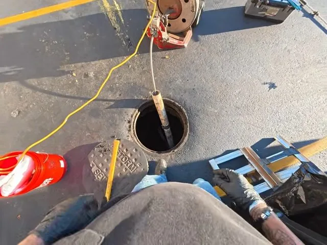 Person in gloves feeding a cable into an open round drain cover on pavement, with a yellow cord and a red bag nearby
