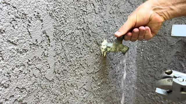 Hand turning a brass hose bib on a stucco wall with water running down from the spout