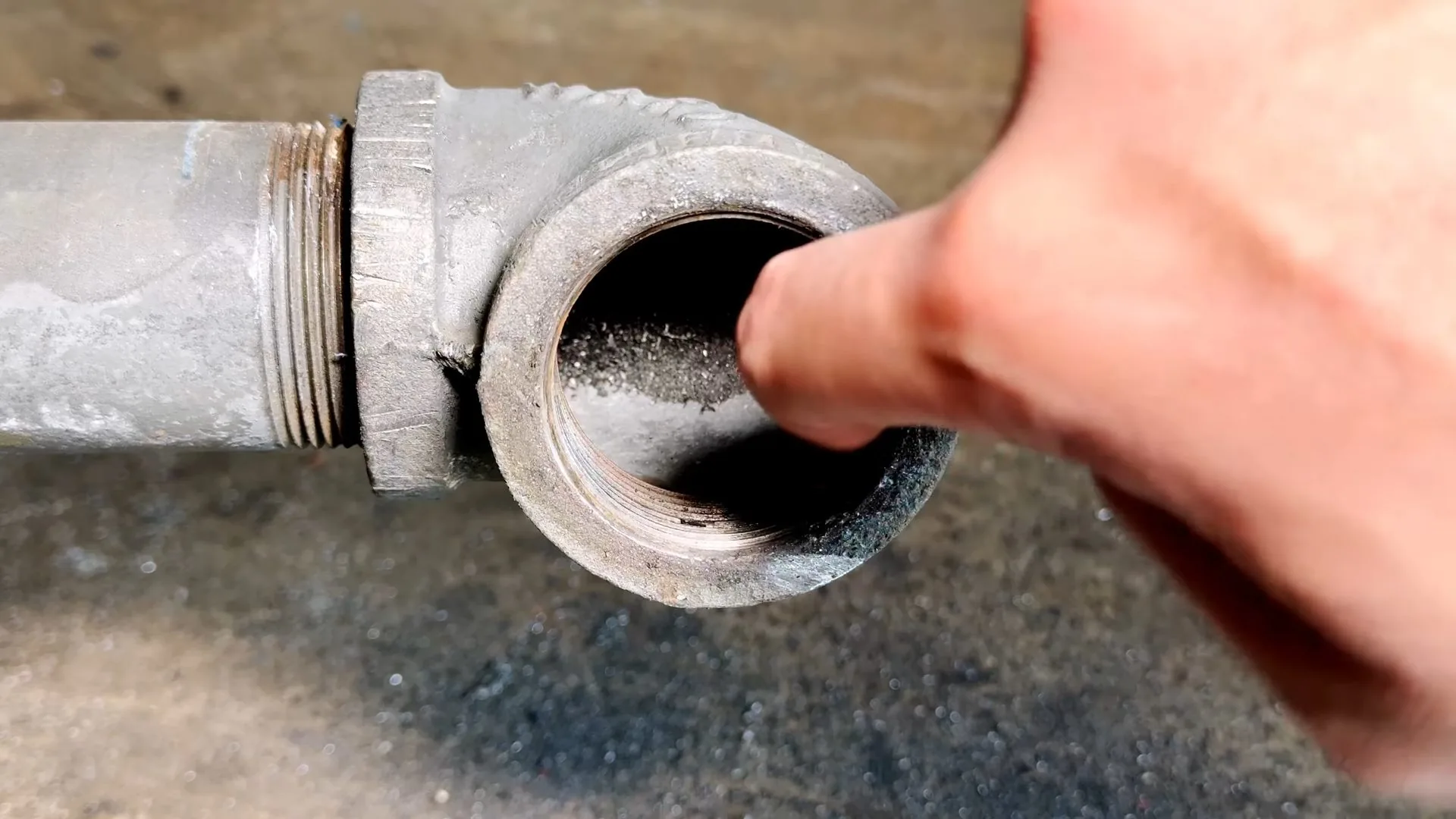 Fingertip inside the open threaded end of a galvanized pipe elbow with gray mineral dust around the rim