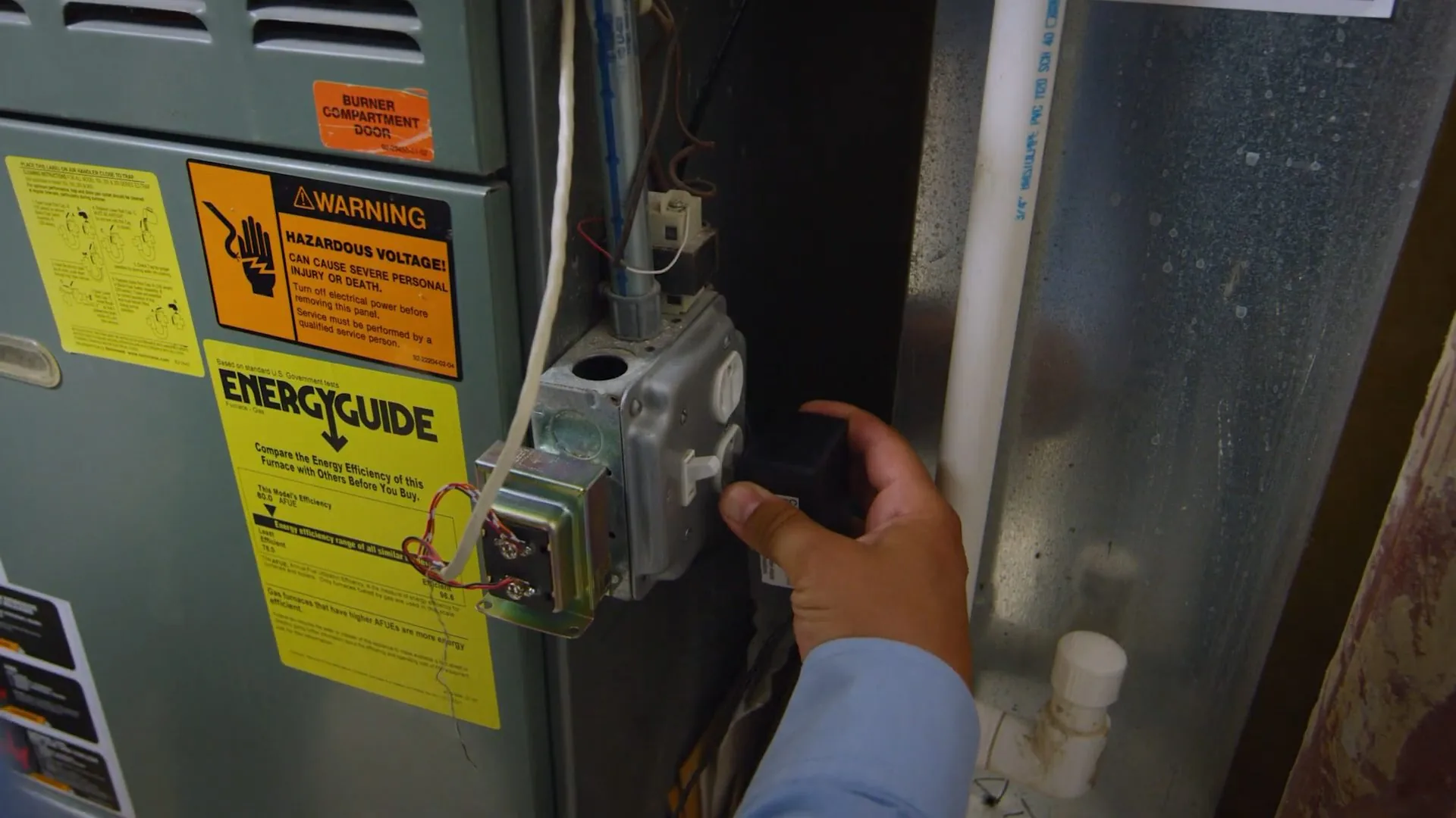 Gas heater cabinet with yellow warning labels and a hand reaching to the gas control valve