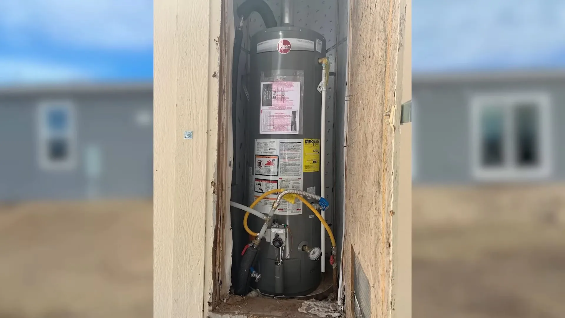 Gas water heater with a yellow gas hose inside a framed wall opening, with a blurred view of houses behind it