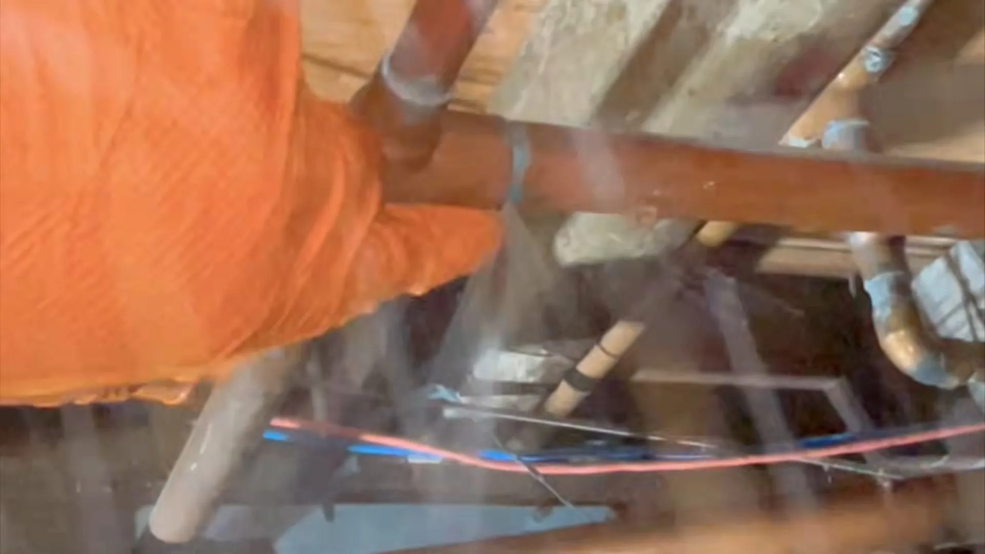 Hand in an orange glove holding a copper pipe inside an opened wall, with water mist in the air around it