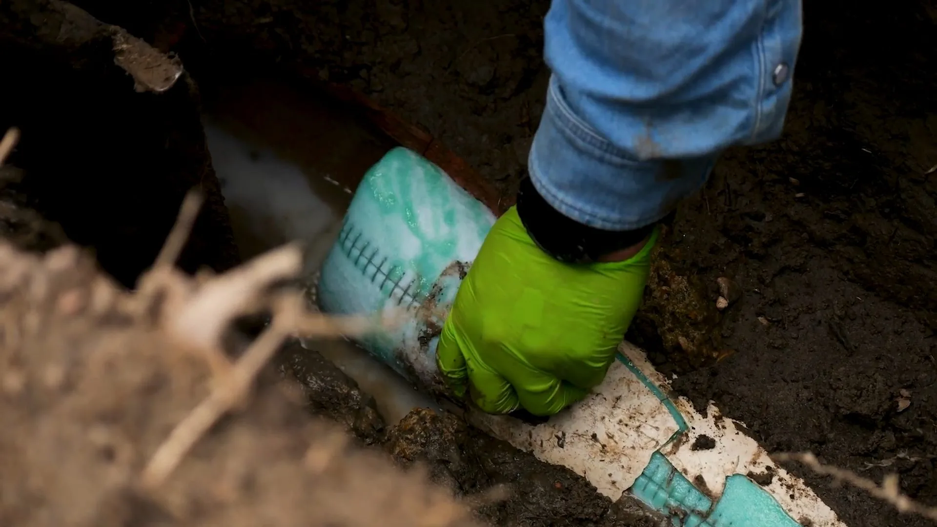 Hand in a green glove on a white pipe with a teal wrap, in a dirt trench with fine roots across it