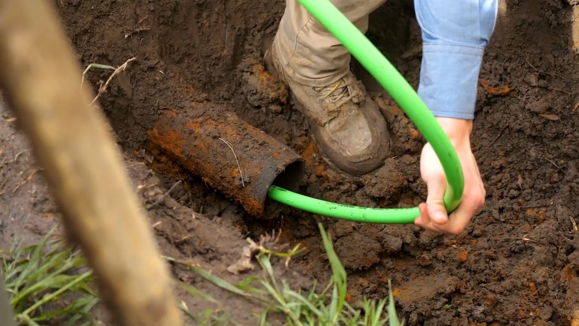 Man in work boots feeding a green hose into a rusted pipe end exposed in a dirt trench