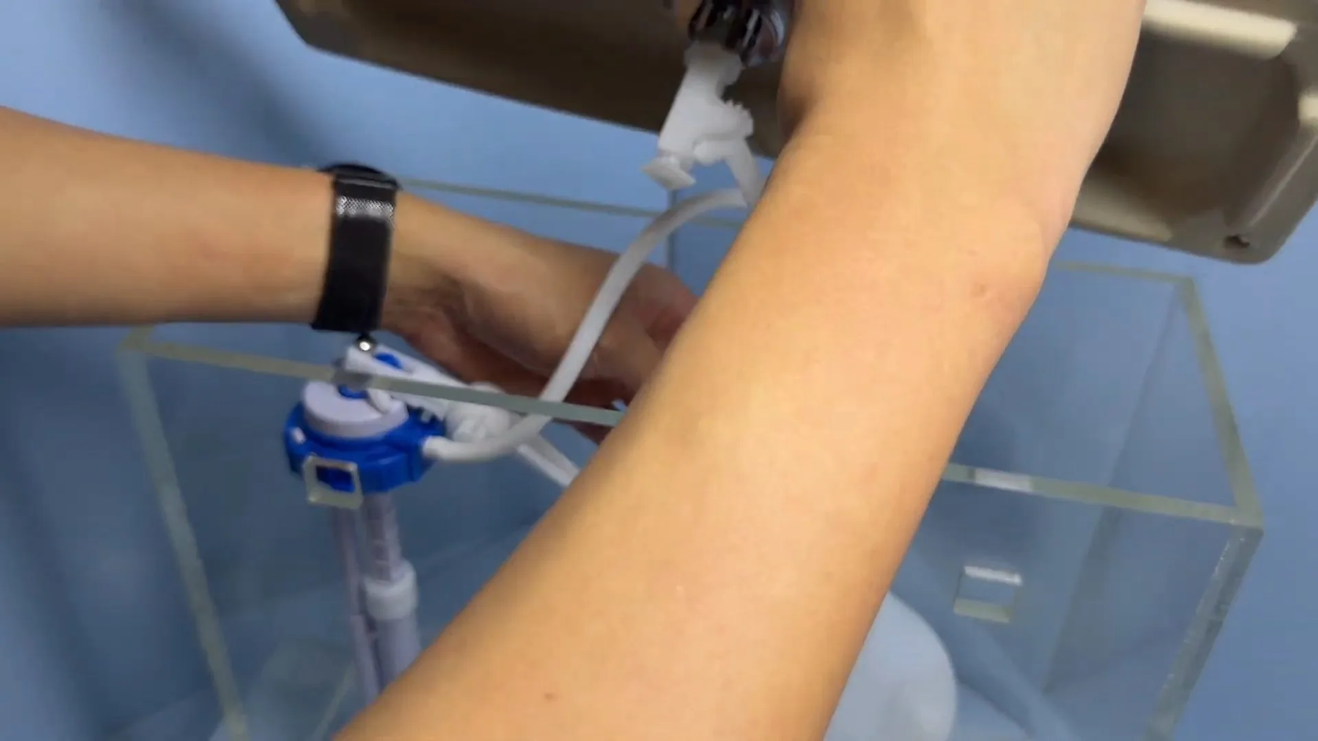 Hand adjusting a blue toilet fill valve inside a clear demonstration tank