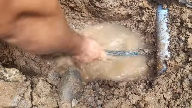 Hand clearing muddy water from around a buried pipe where water is pooling in the dirt
