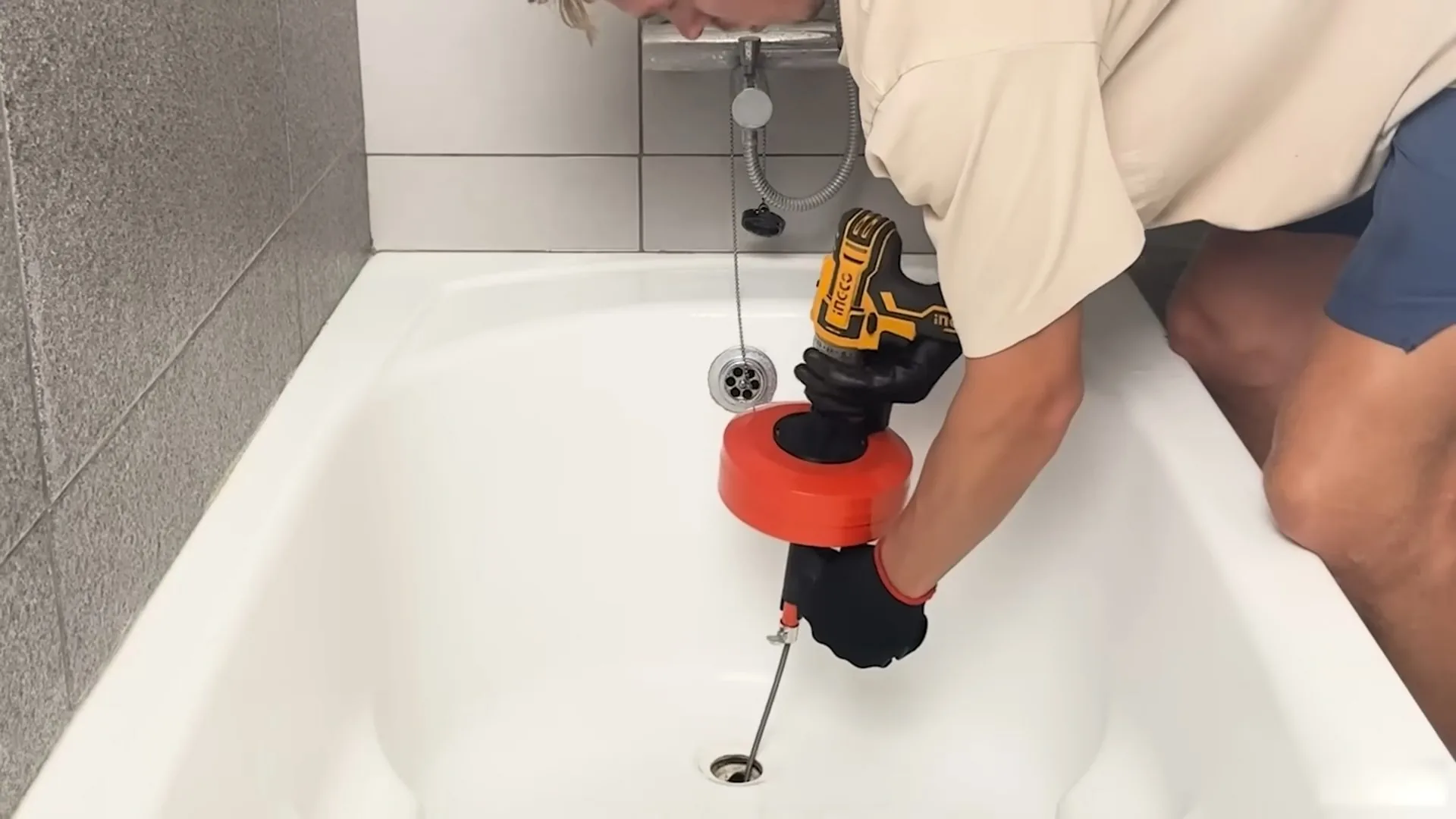 Man in a tan shirt clearing a bathtub drain with a red drain auger turned by a cordless drill