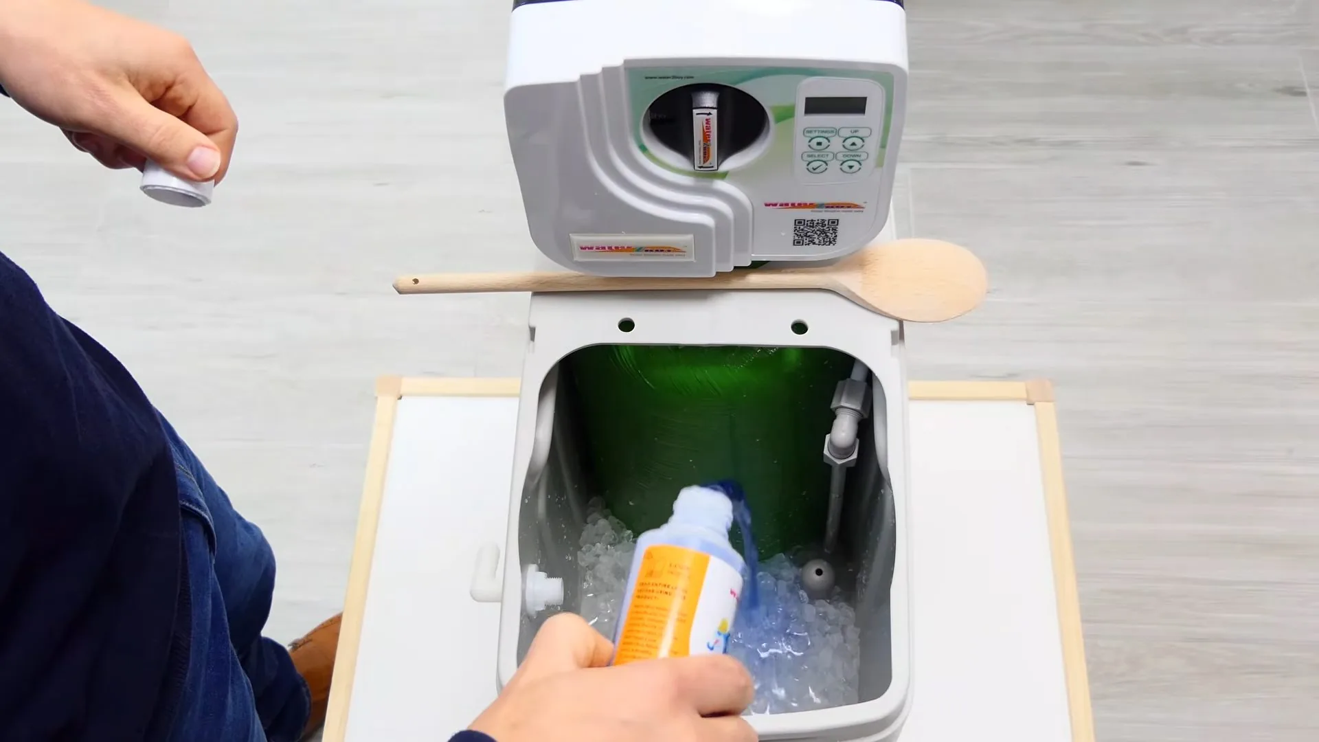 Hand holding an orange bottle over the open brine bin of a water softener with salt inside