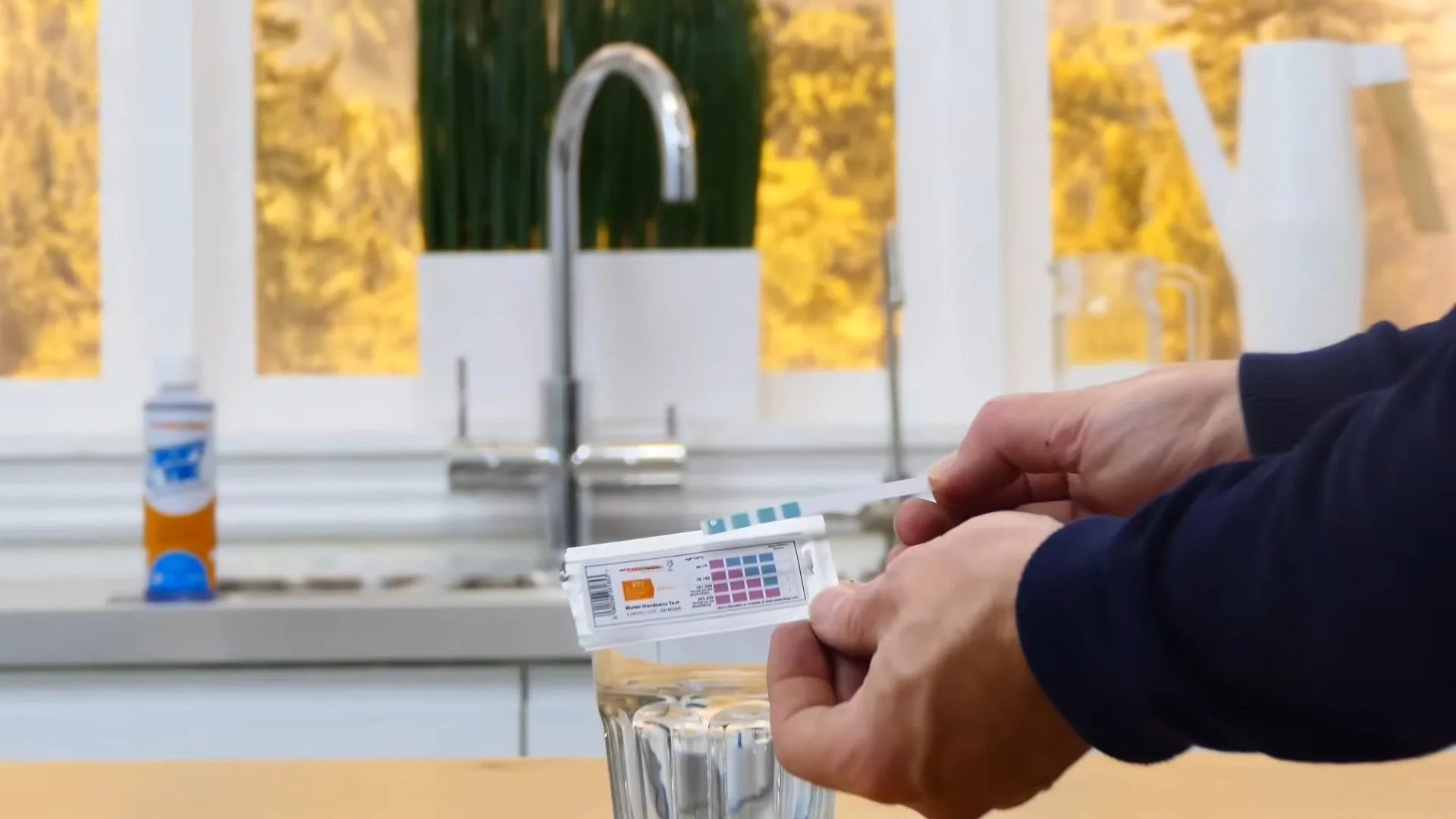 Hands holding a water test strip over a glass of water on a kitchen counter, with a tall faucet behind