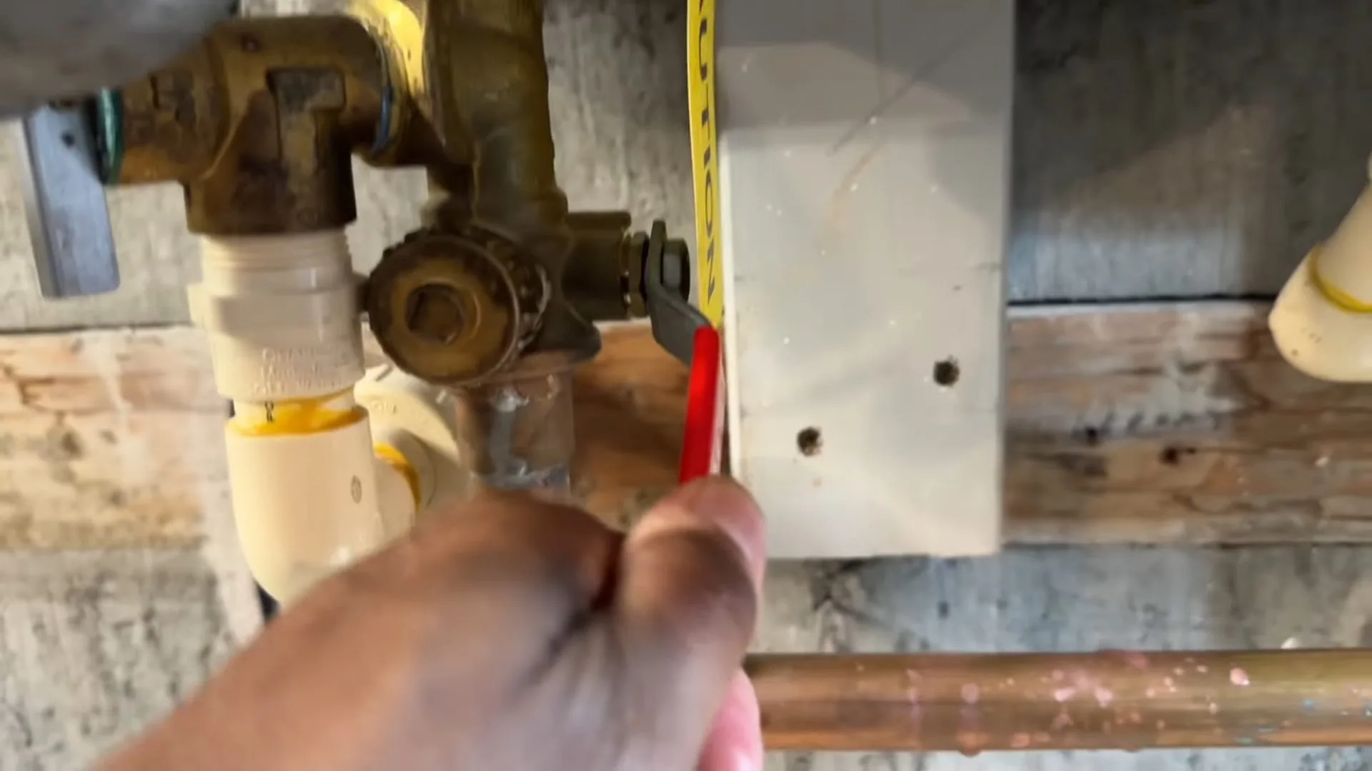 Hand holding a tape measure against copper pipe and a brass valve in a cramped space with white plastic pipes