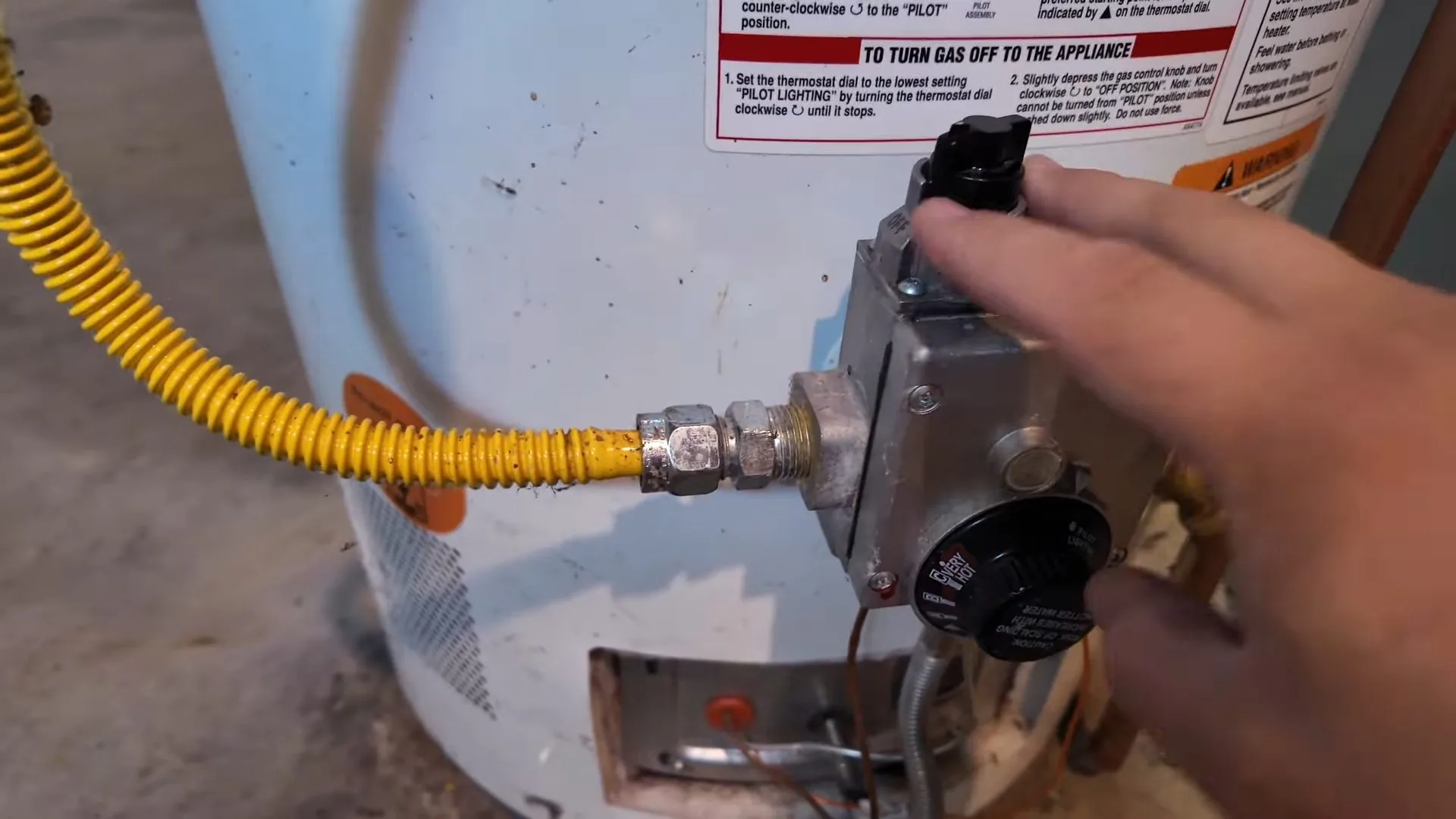 Hand turning the gas control and thermostat on a white water heater with a yellow flexible gas line