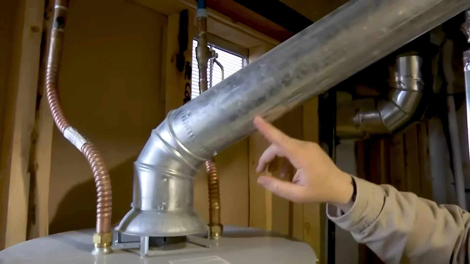 Hand pointing at the metal vent pipe connector on top of a water heater, with flexible gas lines beside it