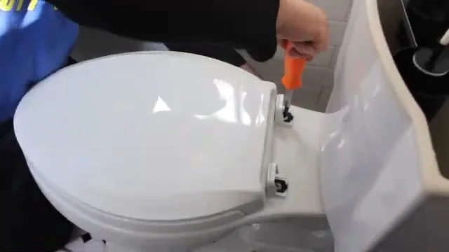 Hand with an orange screwdriver tightening a bolt on the seat hardware of a white toilet
