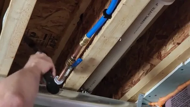 Hand tightening a fitting on blue PEX pipe between wooden ceiling joists, with a red line beside it