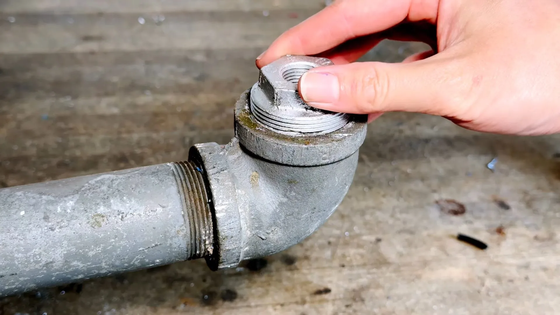Hand unscrewing the square-headed plug from a galvanized pipe elbow lying on a workbench