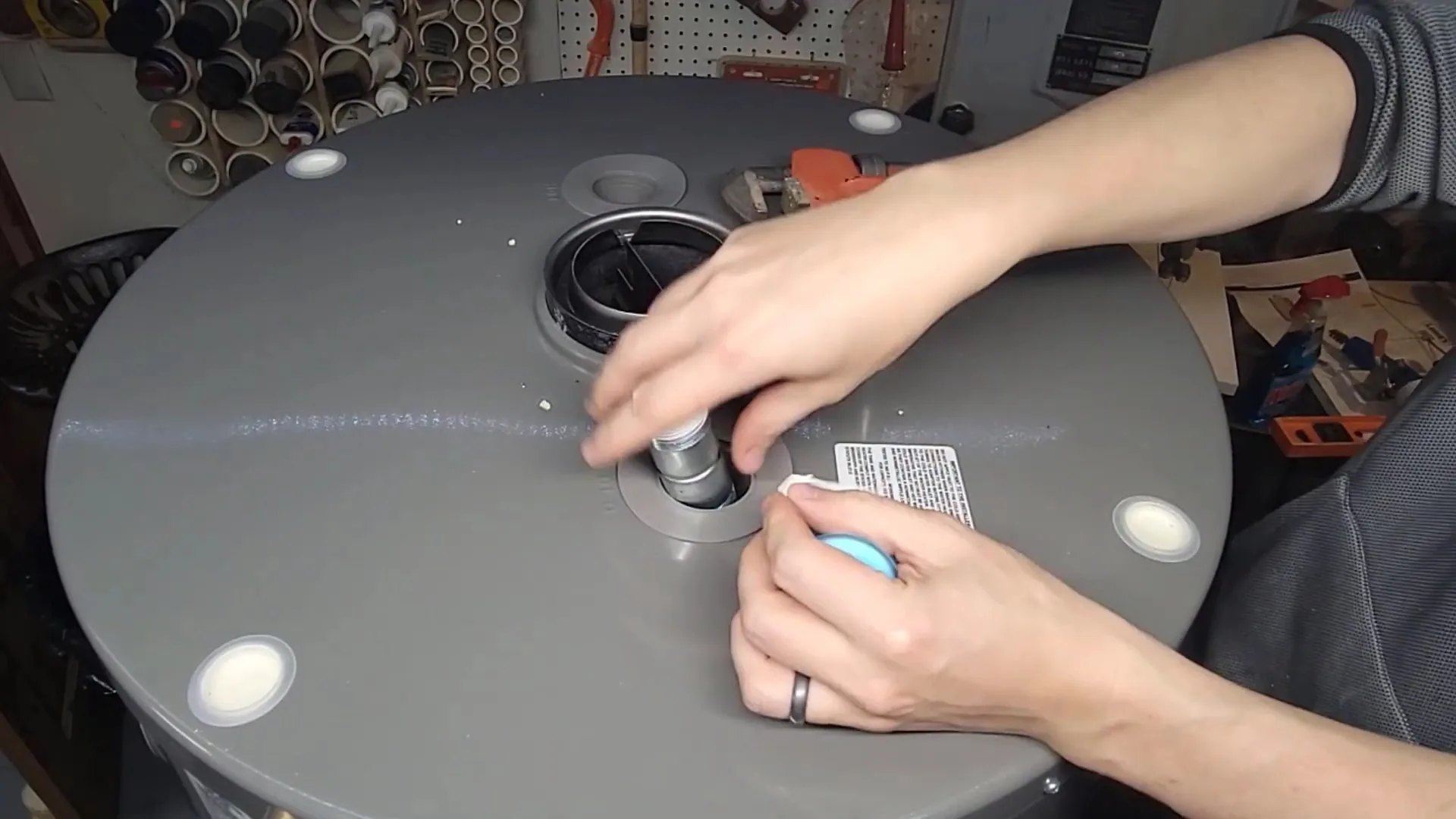 Two hands working at a fitting on the round gray top of a water heater tank