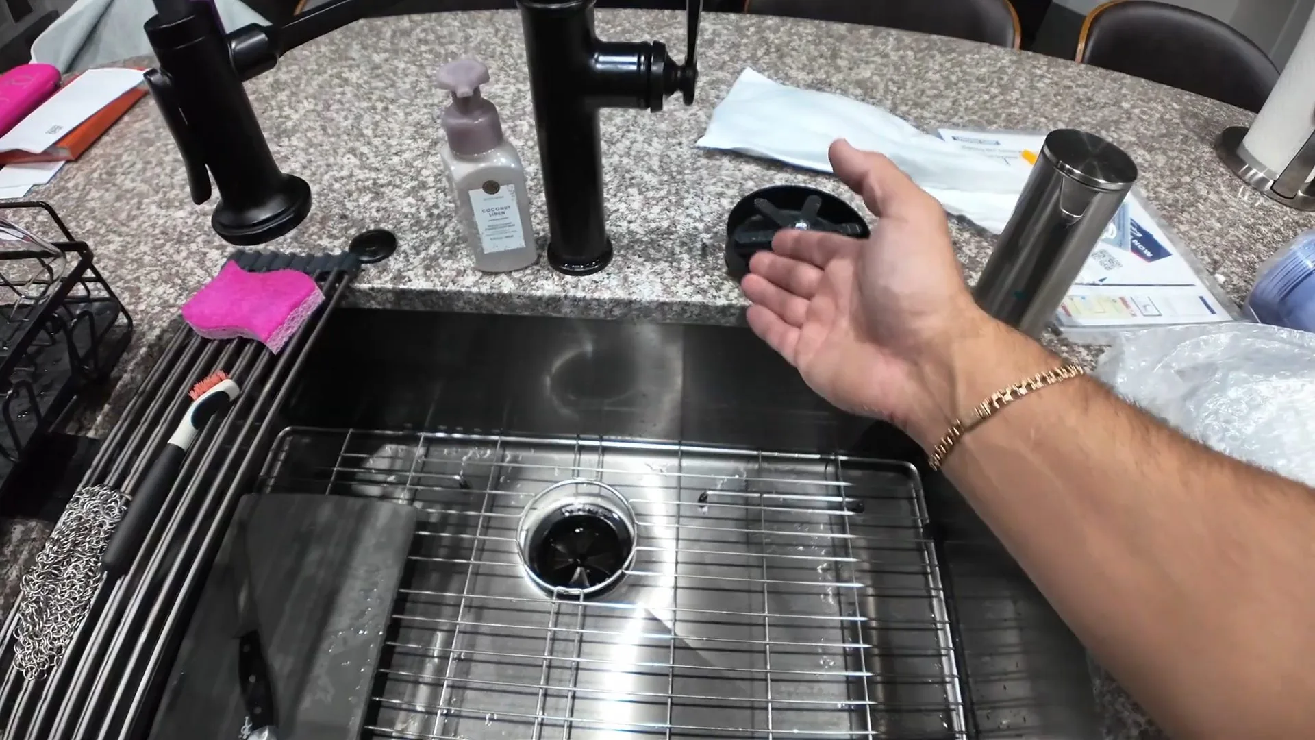 Forearm and hand over a stainless steel sink with a wire grid, a strainer drain and a black faucet