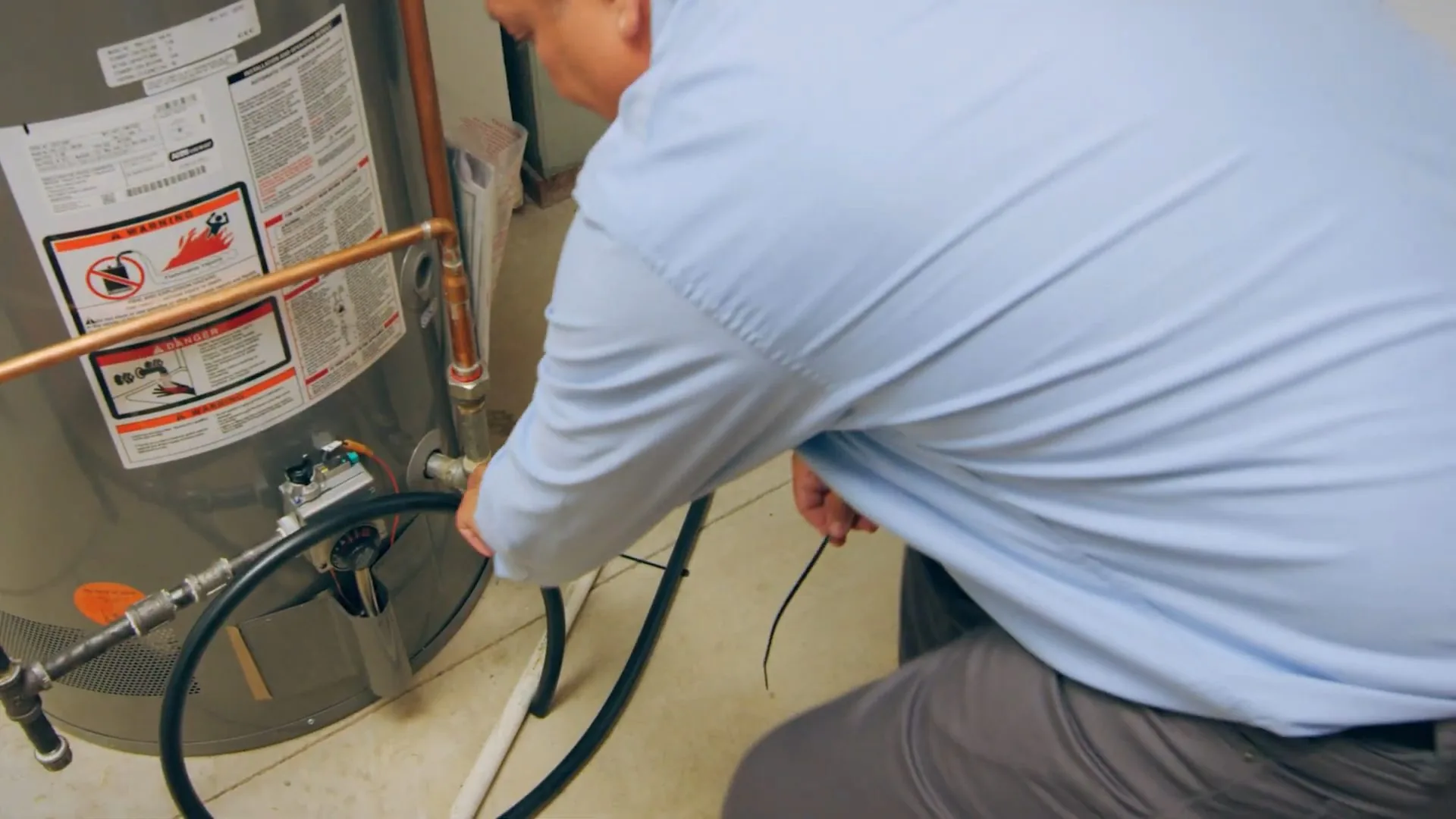Man in a light blue shirt crouched beside a gas water heater working near the base with a hose attached