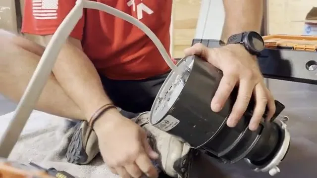 Man in a red shirt lifting a garbage disposal unit toward a sink flange, with a hose beside him