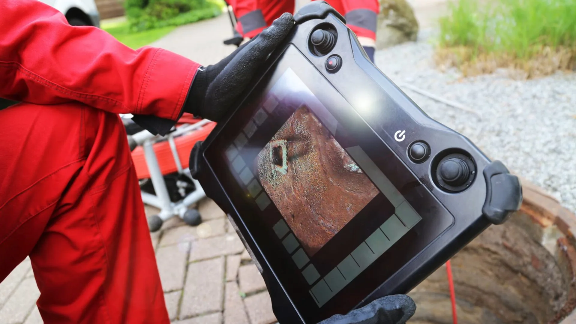 Man in a red jacket holding a rugged handheld camera display beside a round cleanout cover