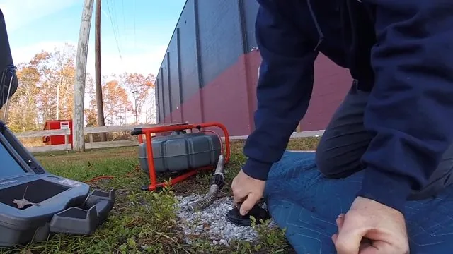 Man kneeling in a grassy yard beside an open cleanout, with a red drain machine and a camera case behind him