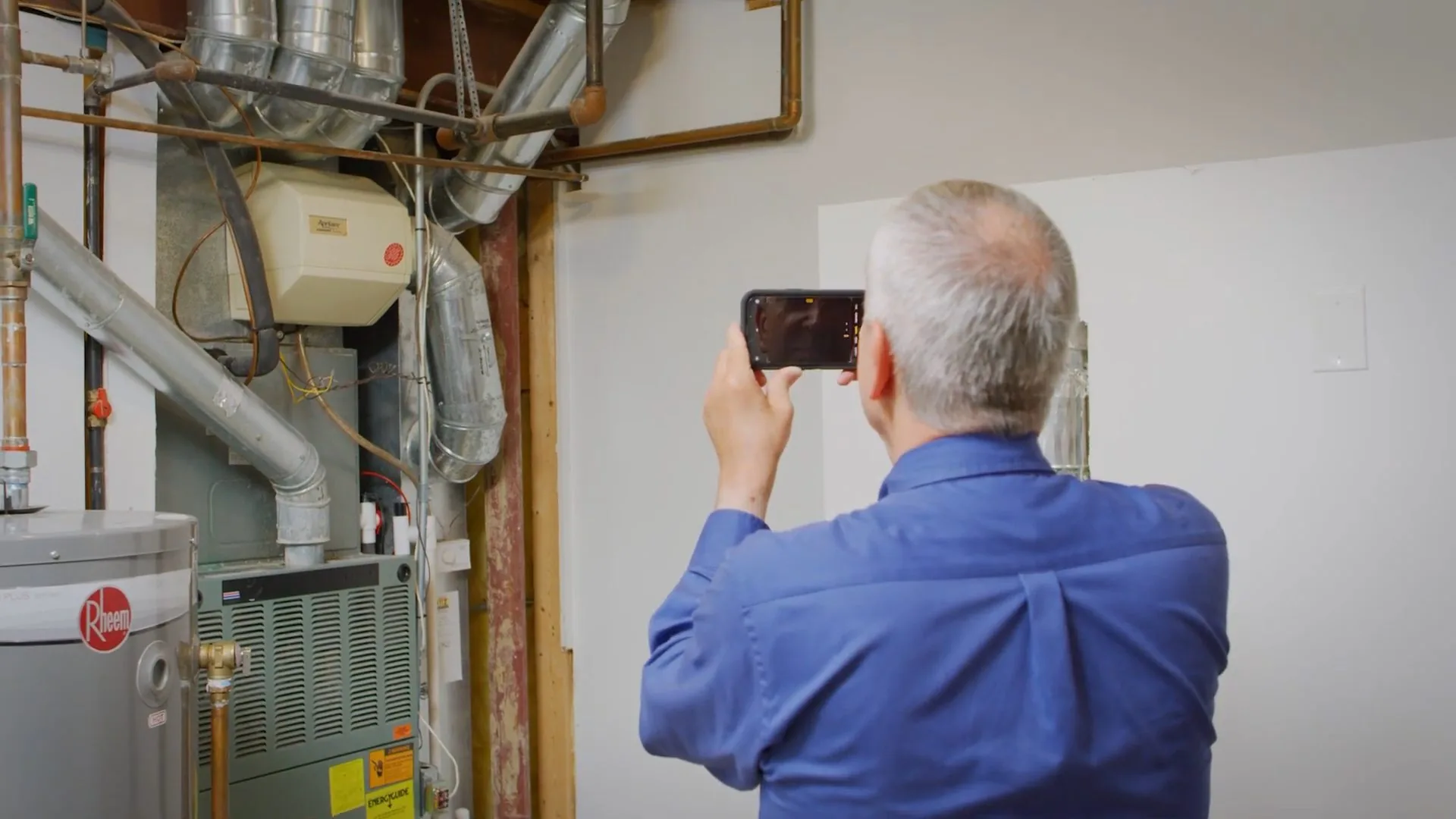Man in a blue shirt taking a phone photo of a water heater and furnace in a utility room
