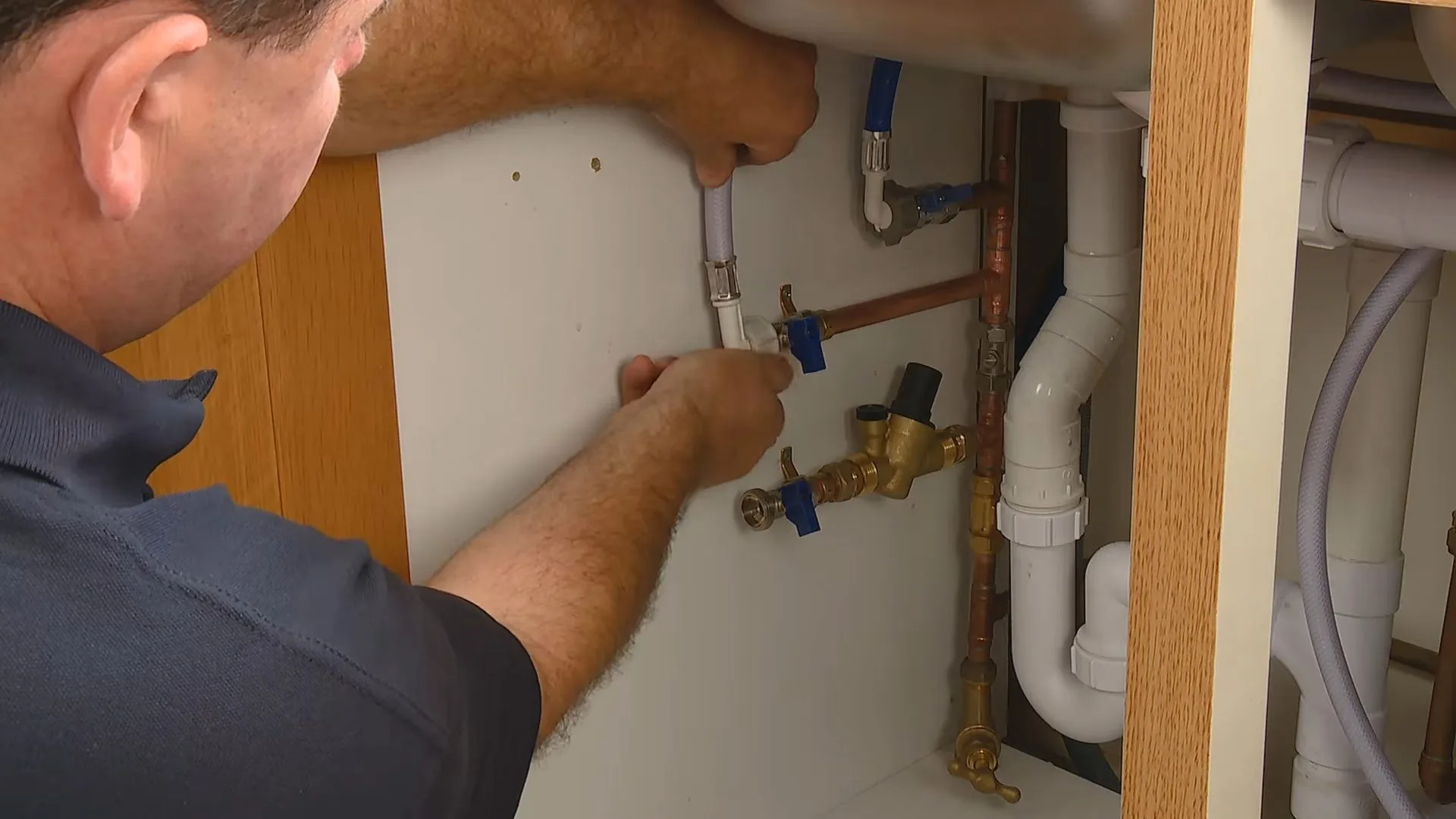 Man in a dark polo reaching under a sink cabinet to two blue-handled shutoff valves on copper stubs