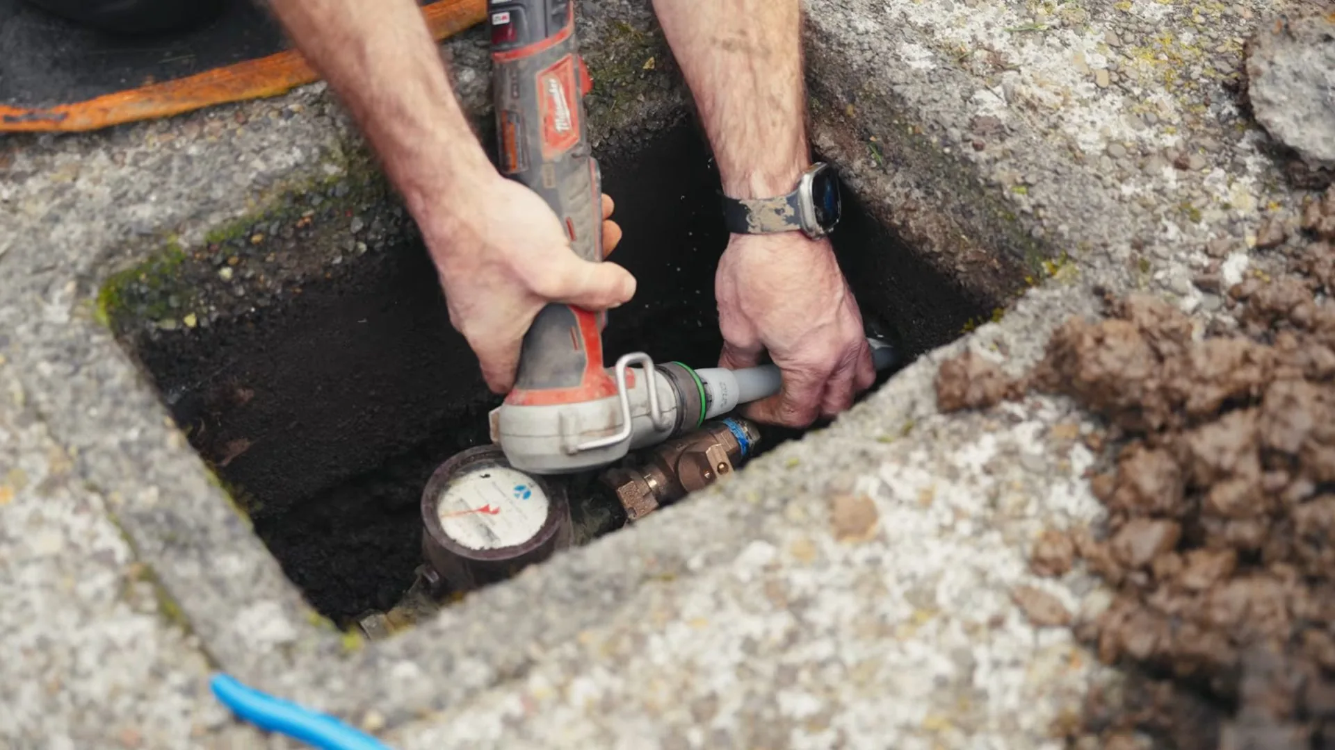 Man holding a power cutting tool over pipe inside an open concrete meter box, with a meter below