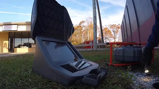 Open black camera control case on grass in front of a brick building, with a red reel behind it