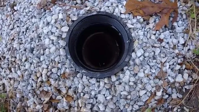 Black pipe opening with its cap off, ringed by white gravel and a few fallen leaves