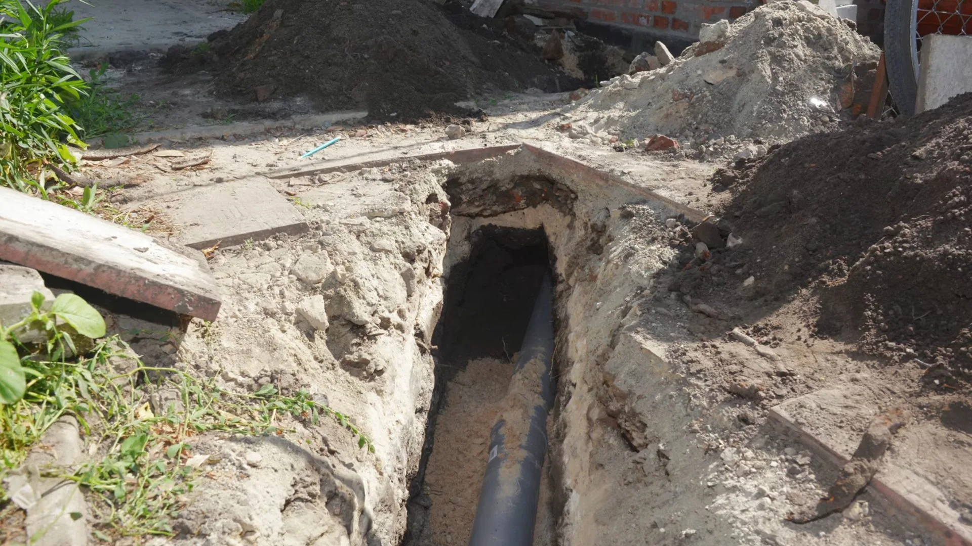 Narrow trench dug beside a building with a dark pipe along the bottom and a mound of sand and soil beside it