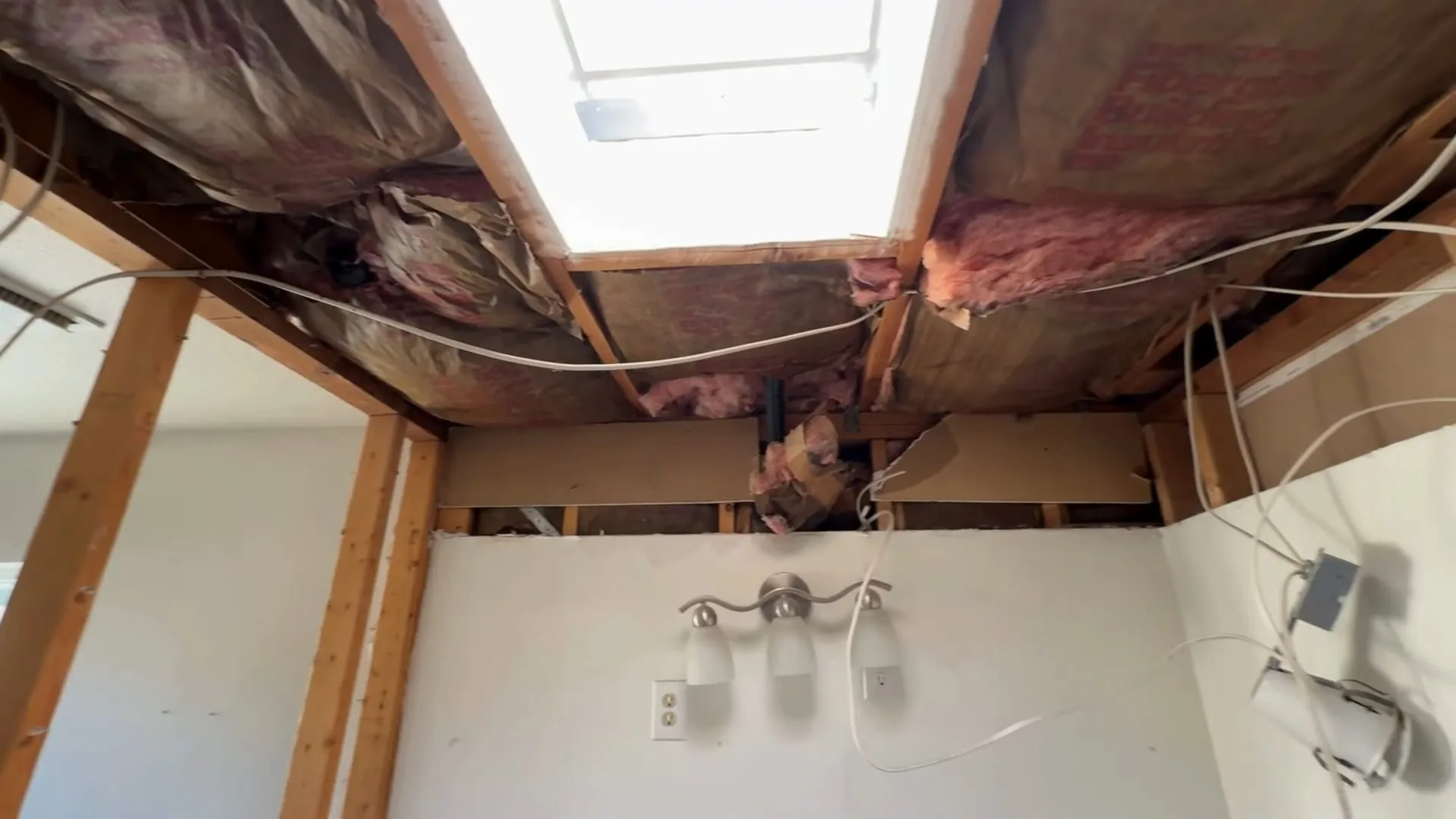 Opened ceiling above a bathroom with pink insulation, exposed joists and wiring