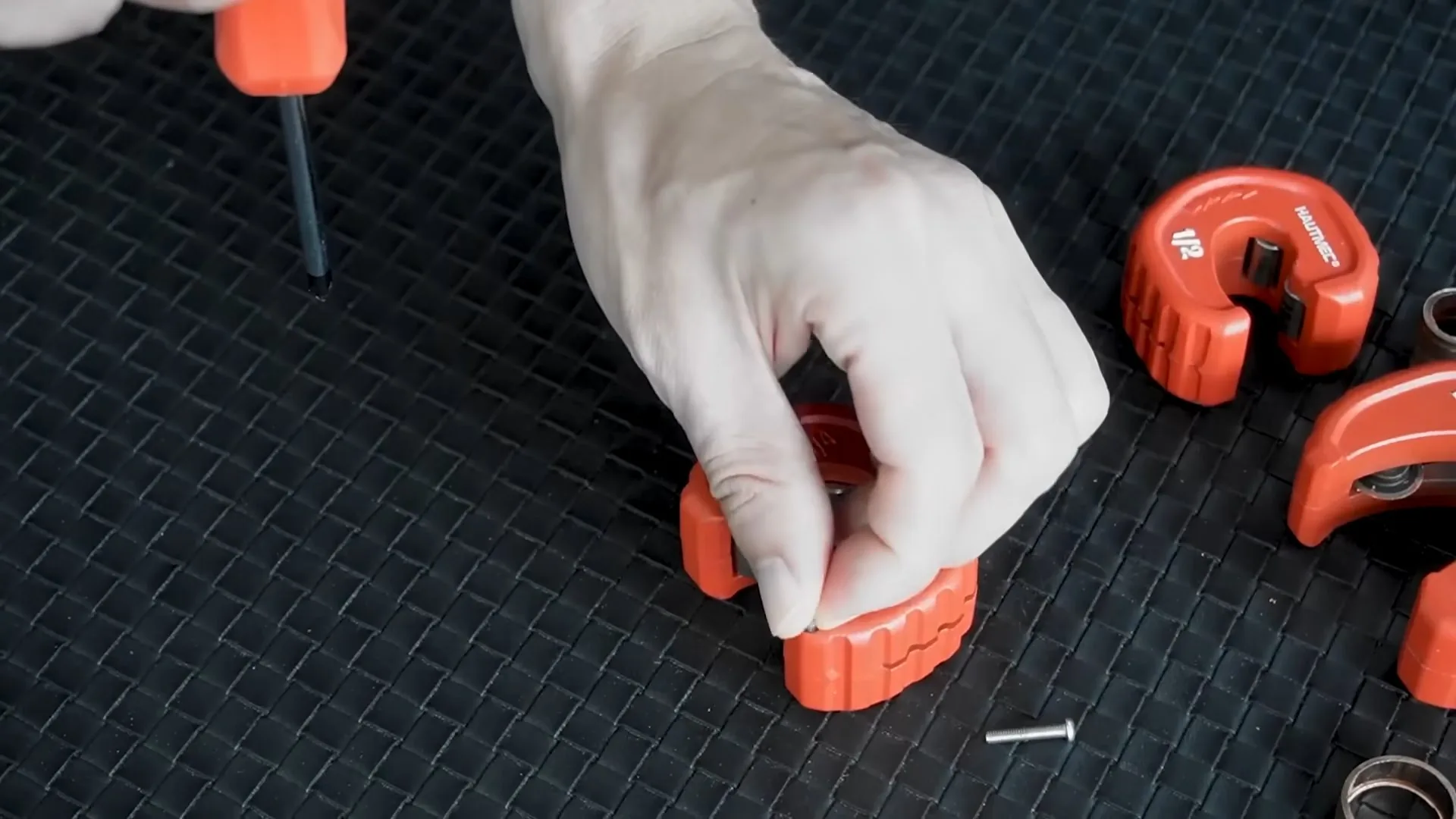 Hand holding one of several orange tubing cutters laid on a black mat beside a screwdriver