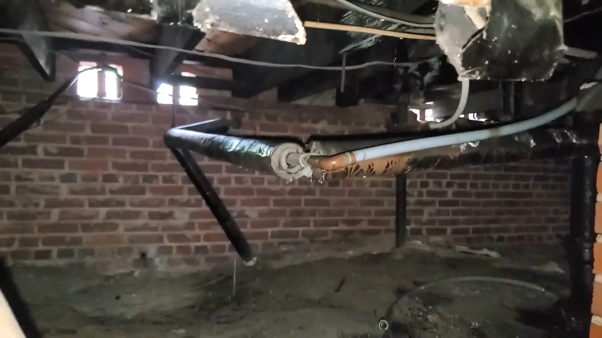 Pipes running beneath a floor in a brick-walled crawlspace with a drop of water falling toward the dirt below