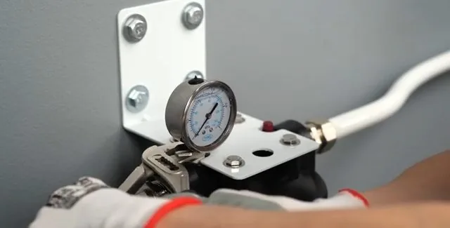 Pressure gauge mounted on a white bracket with a white hose attached, held by gloved hands