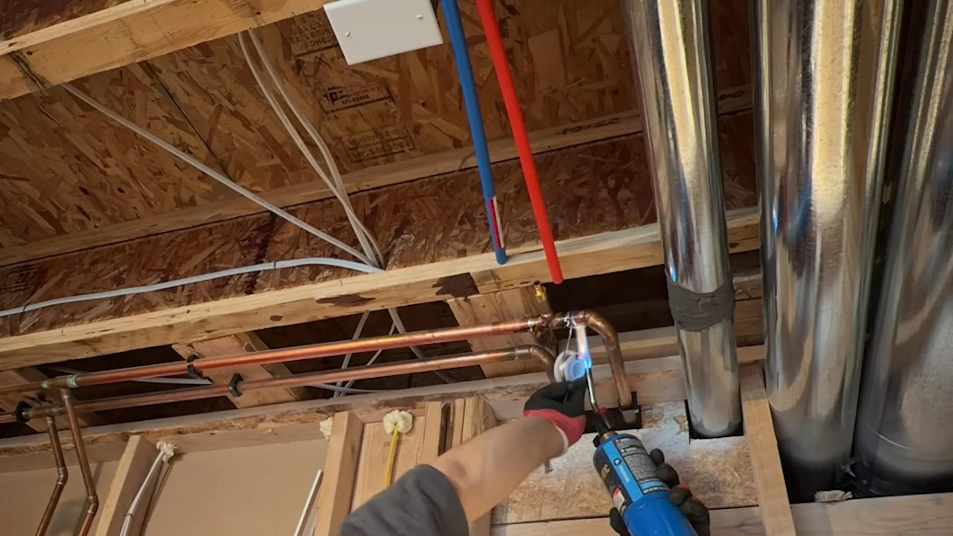 Open ceiling joists with red and blue plastic pipes and a hand holding a tool at a fitting