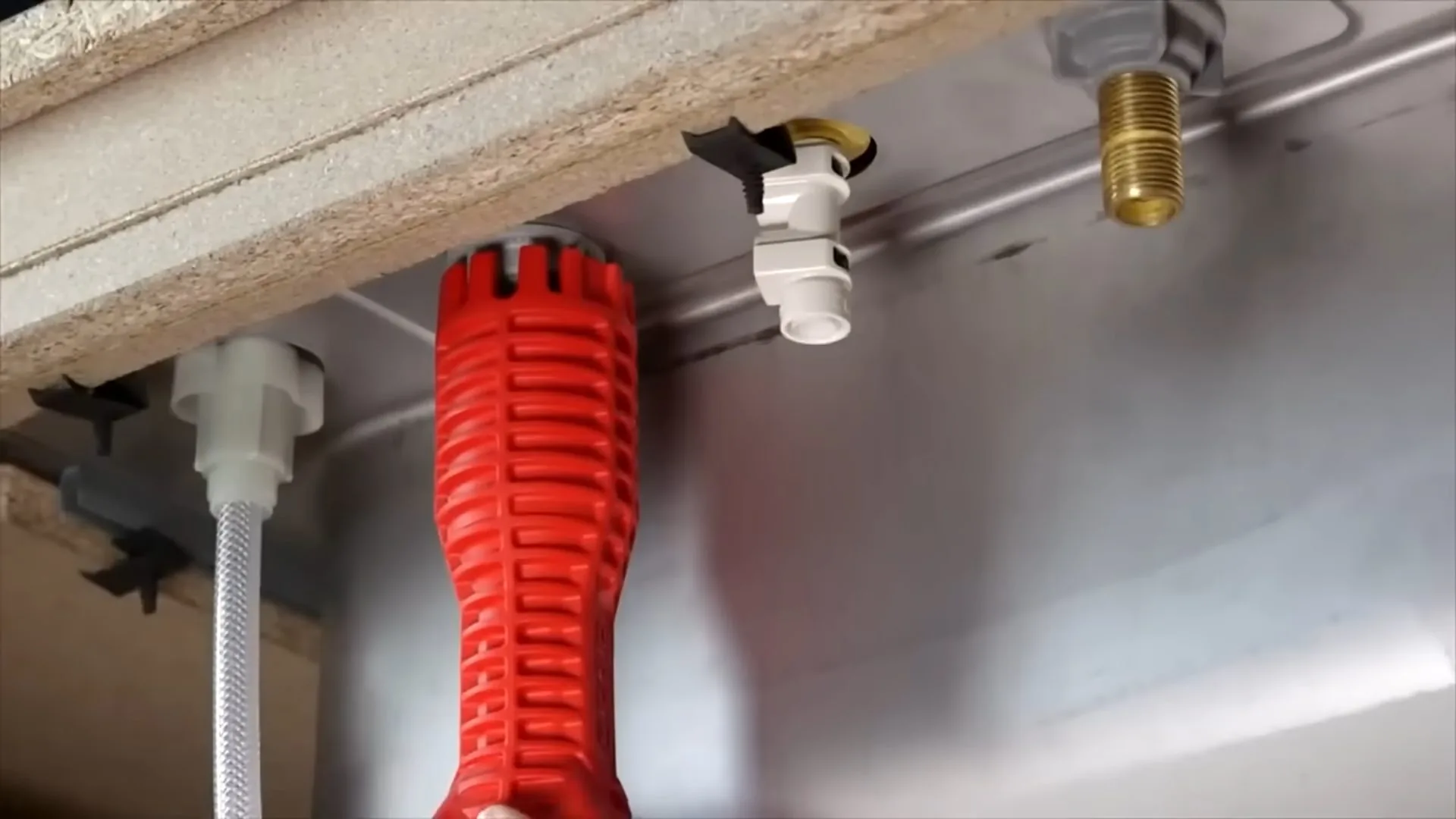 Red basin wrench held up under a kitchen sink beside a white supply line and two shutoff valves