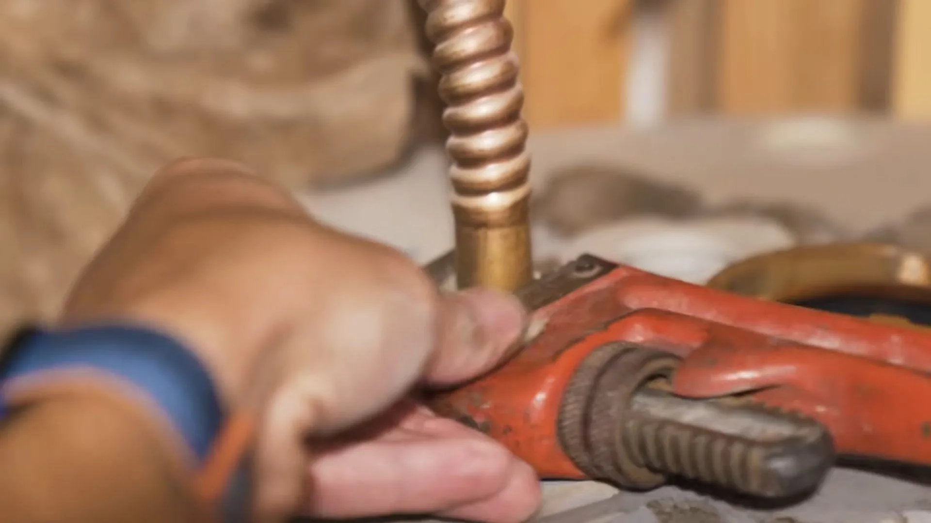 Hand gripping a red pipe wrench on a copper tube beside a corrugated brass fitting