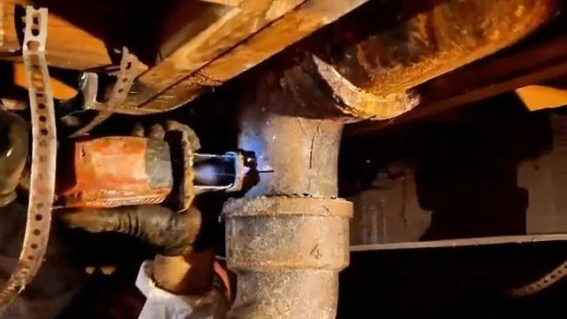 Rust-orange cast iron drain pipe and fitting beneath wooden floor joists, with a metal strap
