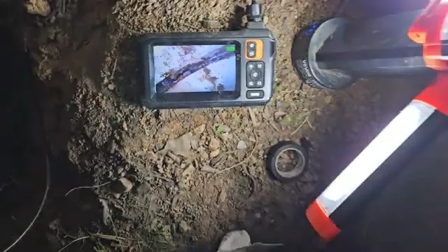 Handheld camera monitor lying on dirt beside an open pipe with a red and white marker
