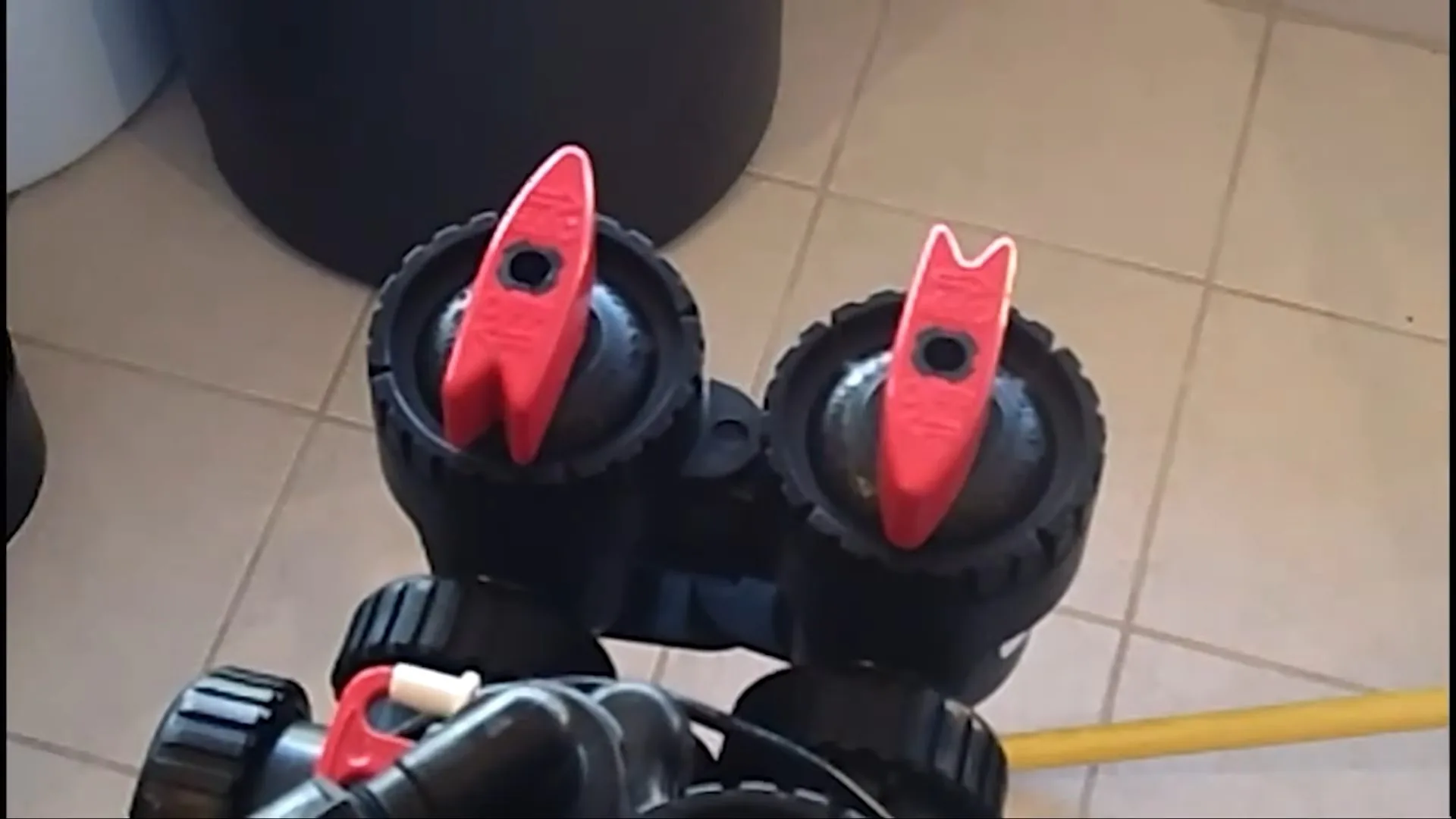 Two red valve handles on a black softener control valve above beige floor tiles