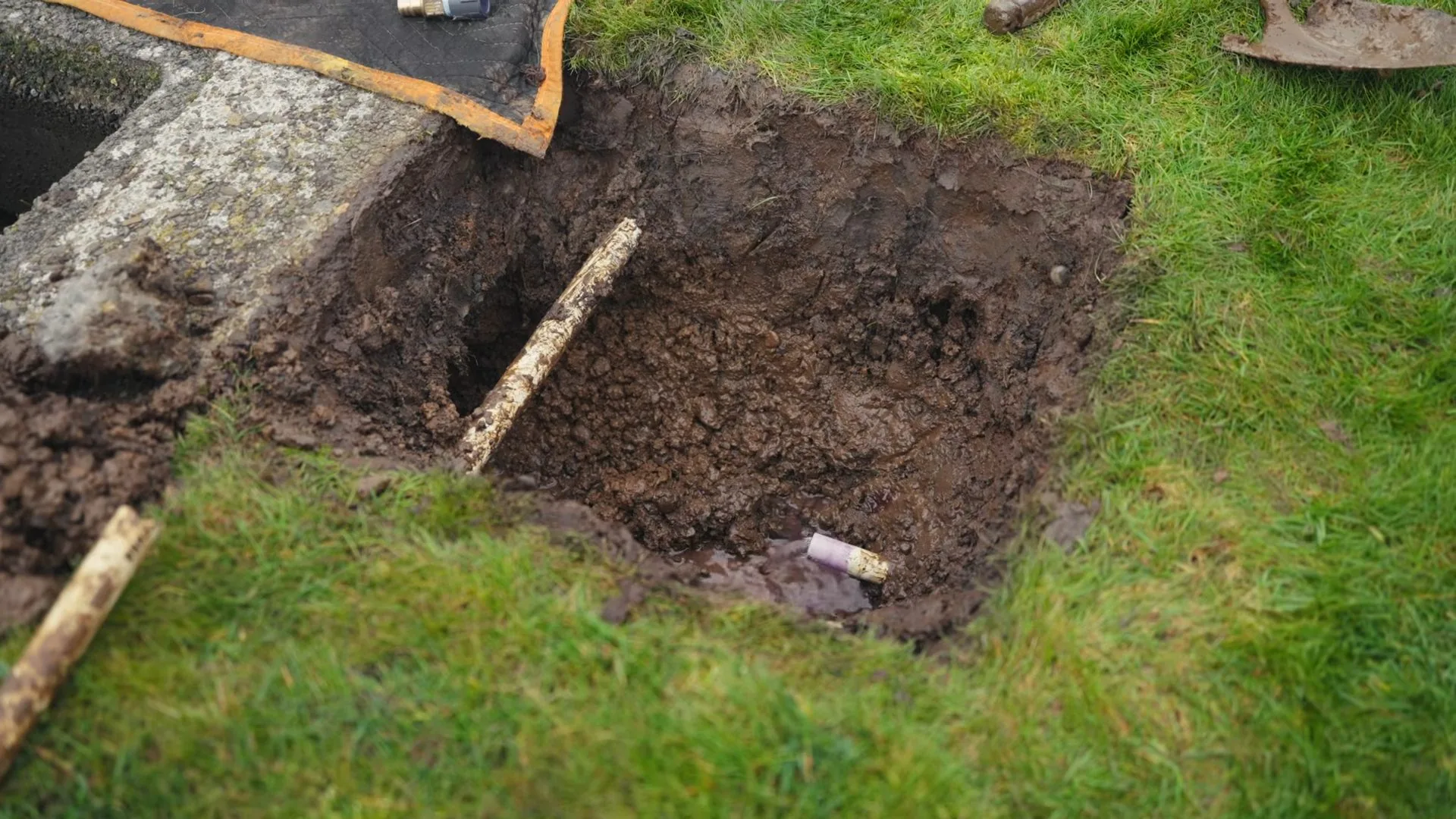 Square hole dug in a lawn with brown soil, a tarp at the edge and a shovel resting nearby