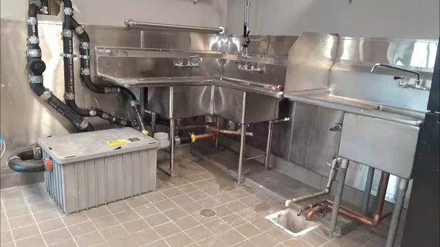 Stainless steel commercial sinks with black drain pipes, copper lines and a gray grease trap on a tile floor