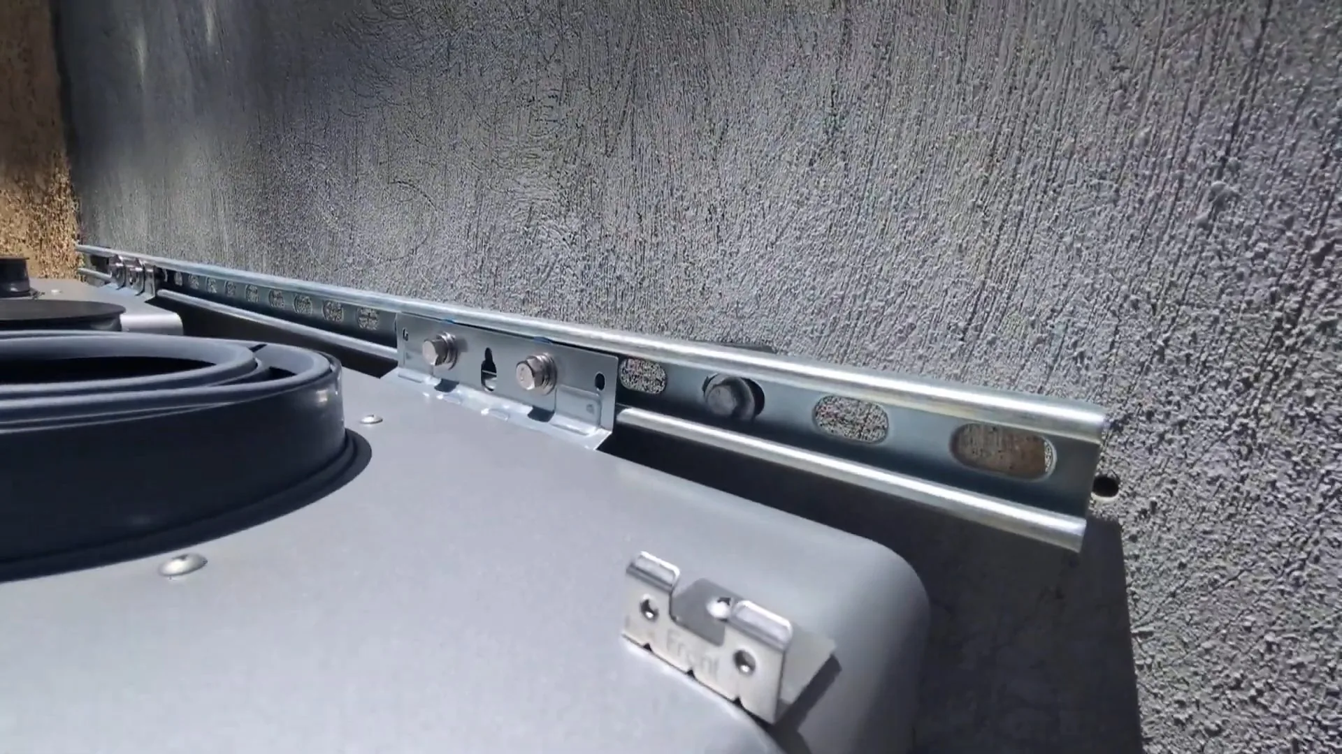 Metal mounting rail and bracket on the top edge of a gray tankless water heater against a dark textured wall