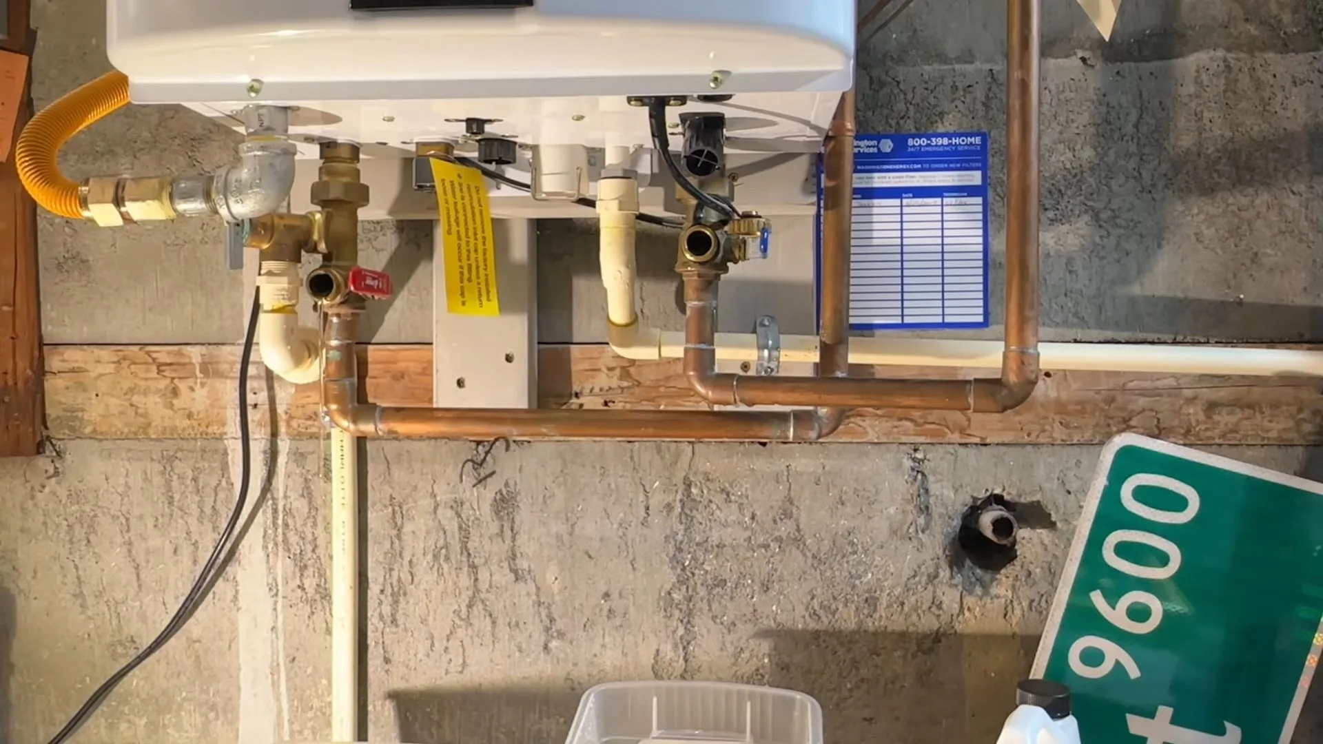 Underside of a tankless water heater with copper pipes, a yellow gas hose, shutoff valves and a service tag, with a green street sign in frame