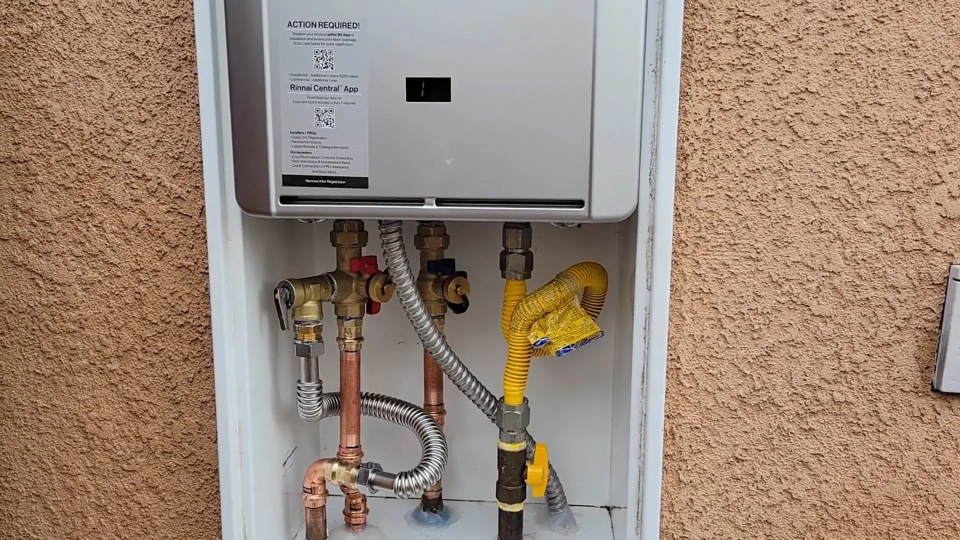 Wall-mounted tankless water heater in a recessed white cabinet on a stucco wall, with copper, flexible and yellow gas pipes below